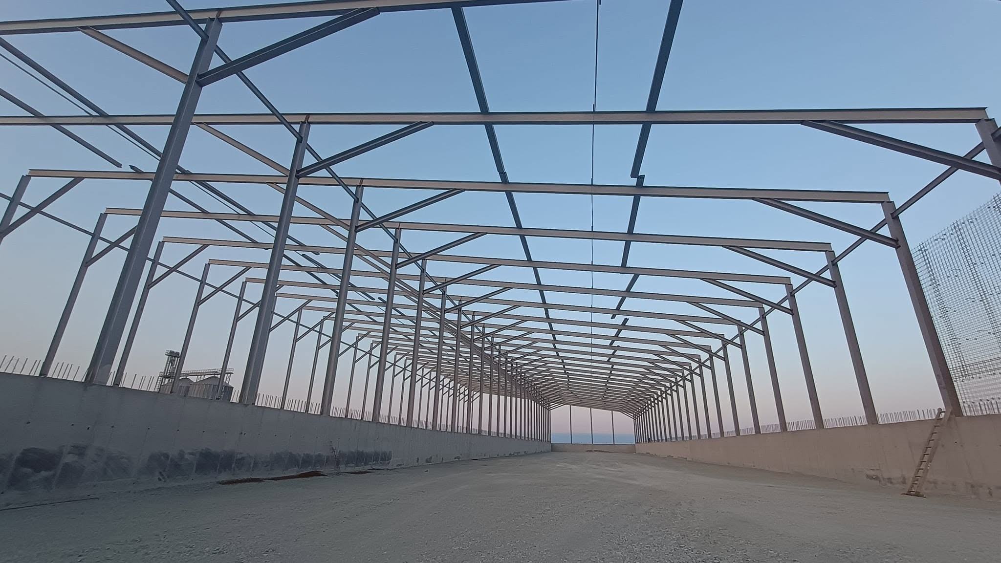 Perspectivă lungă printr-o hală metalică în construcție, cu pereți din beton