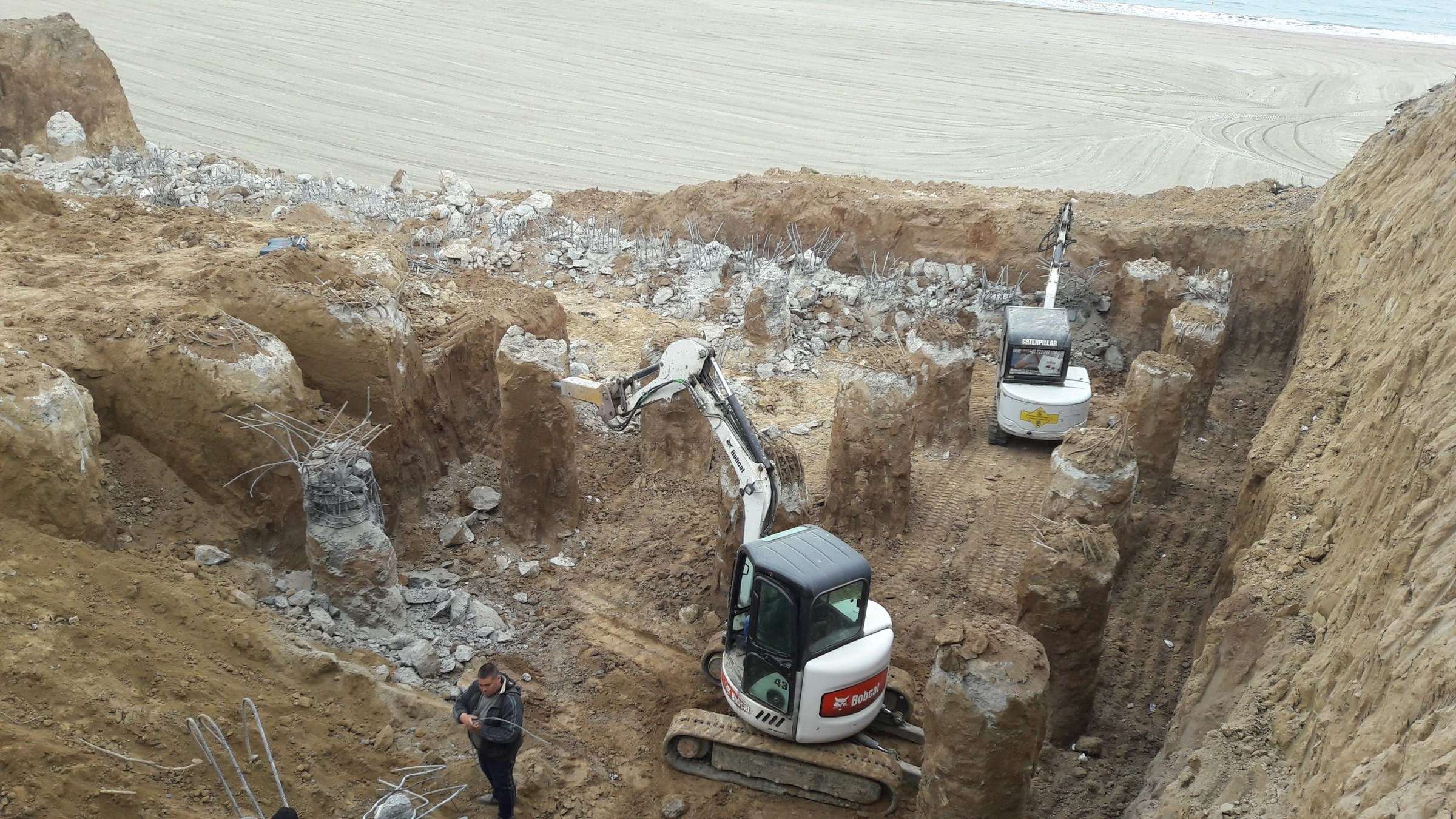Piloți forați cu armătură, două miniexcavatoare în groapa de fundație, plaja și marea în fundal