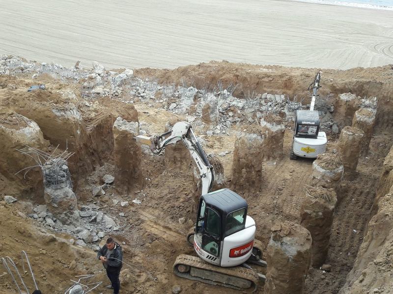 Piloți forați cu armătură, două miniexcavatoare în groapa de fundație, plaja și marea în fundal